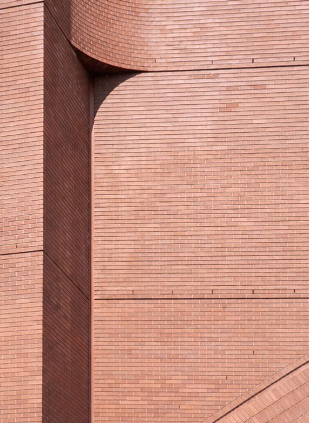 Close-up architectural detail of a red-pink brick building facade. The image shows tightly coursed horizontal brickwork, a deep vertical recess, and a curved projecting brick form creating a strong shadow and sculptural geometric composition. No interior space is visible.