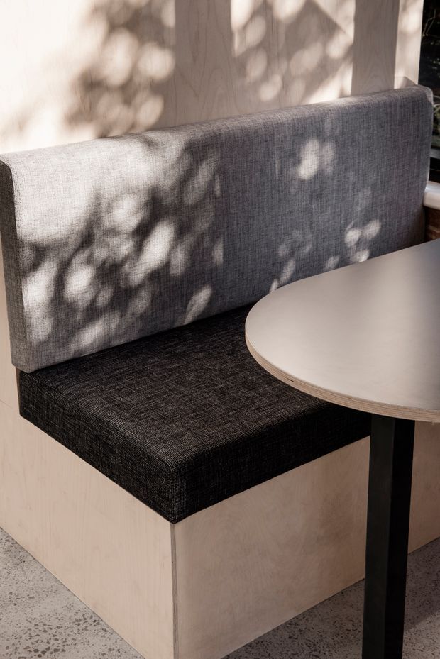 Close-up interior view of a built-in booth seat with a charcoal textured upholstered bench cushion and backrest, paired with a round light neutral tabletop on a black pedestal base. Pale timber wall and base panels surround the seating, with dappled sunlight creating organic shadows. The floor appears to be a speckled terrazzo or concrete finish.