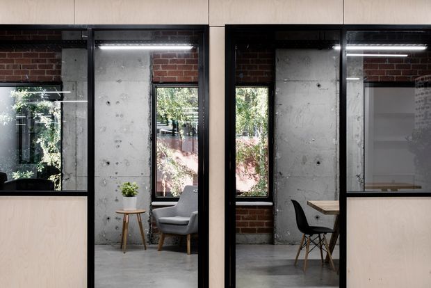 Interior view of a compact industrial-style workspace or studio divided by black-framed glazed partitions and pale timber panels. Exposed red brick, raw concrete walls and polished concrete flooring create a strong material contrast, while windows bring in greenery and daylight. A small desk and chair are visible in one bay, with a lounge chair and side table in another.