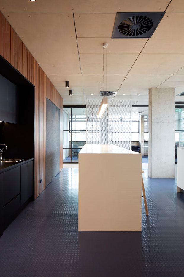A contemporary open-plan commercial interior with a central white service island or counter, black built-in cabinetry, timber slatted wall panelling, exposed concrete columns, glazed partitions, and suspended linear lighting. The ceiling features light timber panels with visible mechanical ventilation, while the dark textured floor creates strong contrast.