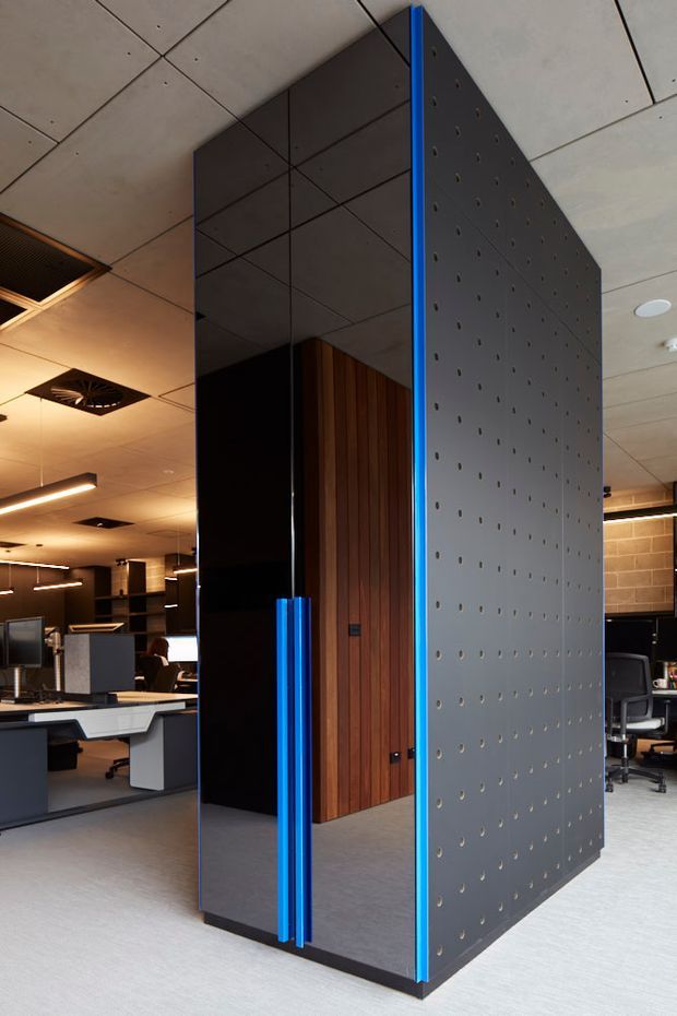 Interior view of a contemporary open-plan office with workstations visible around a prominent freestanding partition or enclosed meeting pod. The structure combines dark reflective panels, warm vertical timber cladding, a perforated grey metal wall, and vivid blue vertical trim and door pulls. Suspended linear lighting, acoustic ceiling panels, and exposed office furniture reinforce the commercial workplace setting.