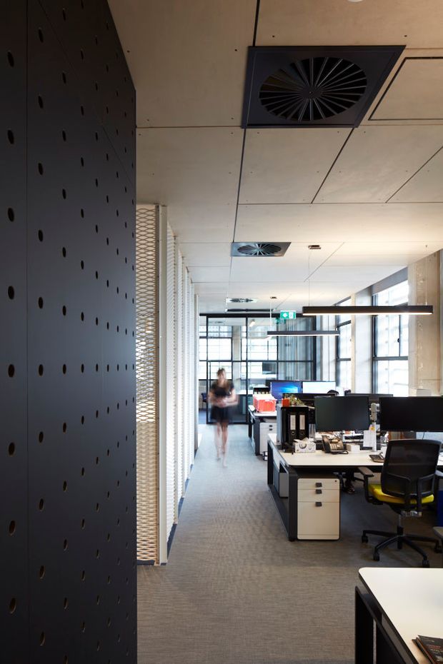 A contemporary open-plan office interior viewed down a central circulation aisle. The space features exposed suspended ceiling panels with integrated circular air vents, linear pendant lighting, carpeted flooring, large glazed windows, black perforated feature wall panels, workstation desks with computer monitors, filing drawers, and office chairs. A glazed meeting or work area is visible in the background.