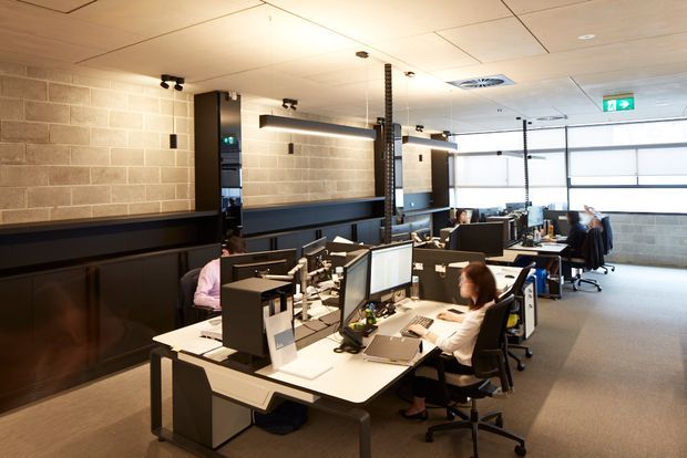 Open-plan commercial office with rows of workstations, computer monitors, task chairs, and employees. The space features exposed concrete block walls, dark built-in cabinetry or wall panelling, suspended acoustic ceiling panels, linear pendant lighting, and large glazed roller doors or windows along the far wall.
