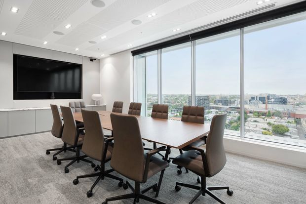 Bright contemporary boardroom with a large rectangular wood meeting table surrounded by upholstered swivel chairs. A wall-mounted display and integrated cabinetry occupy one side, while full-height windows provide expansive city views. The room has a white acoustic ceiling with recessed lighting and a neutral carpeted floor.