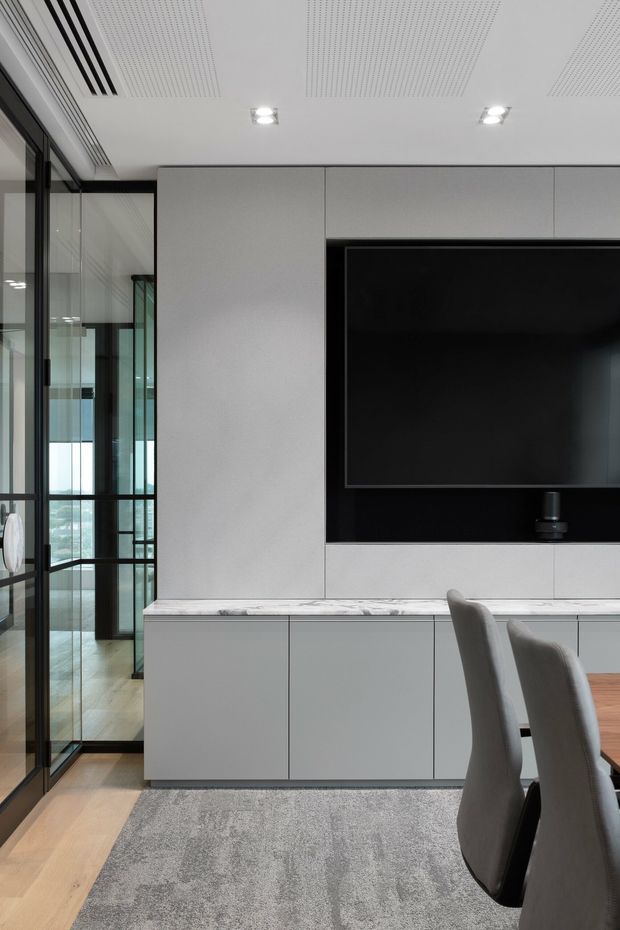 Contemporary open-plan dining and living area featuring a full-height white built-in media wall with a large recessed black television, integrated low cabinetry, and a pale stone benchtop. Dark-framed glazed doors and partitions line the left side, opening toward a bright adjoining space. Grey upholstered dining chairs, a light timber floor, and a large grey rug create a restrained, refined interior. The ceiling has recessed downlights and perforated acoustic panels.