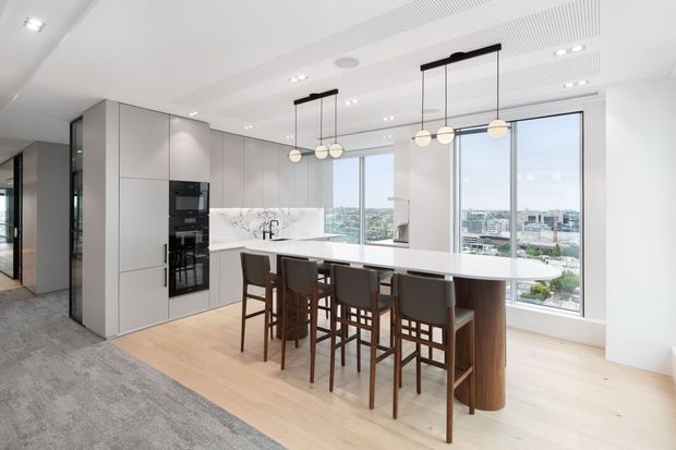 Bright contemporary open-plan kitchen with a large white island and integrated dining seating, floor-to-ceiling windows overlooking an urban cityscape, pale timber flooring, streamlined grey cabinetry, and linear pendant lighting. A living area carpet is visible in the foreground.