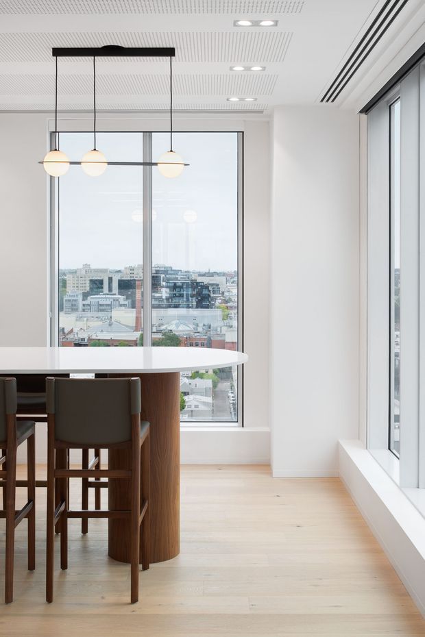 Bright contemporary dining area in a high-rise setting, featuring a rounded white dining table with a dark timber pedestal base, timber-framed upholstered bar chairs, a linear pendant with three spherical globes, pale timber flooring, white walls, a perforated acoustic ceiling, and expansive floor-to-ceiling windows overlooking an urban cityscape.