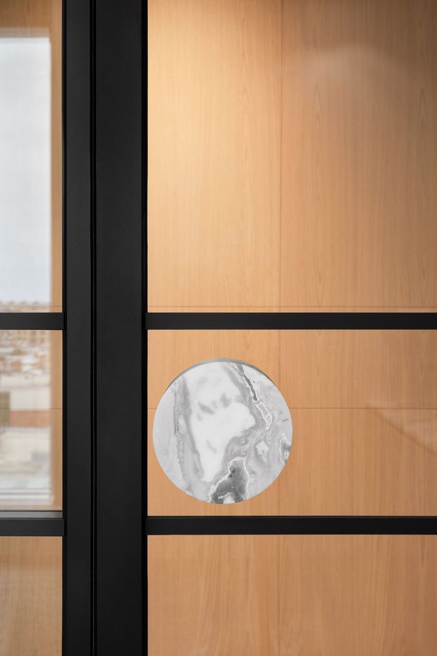 Close-up architectural detail of a timber-panelled door or partition with bold black metal framing and horizontal mullions. A circular grey-white marble handle or decorative inset provides a contrasting feature. Glazing at the left reveals a softly blurred interior beyond.