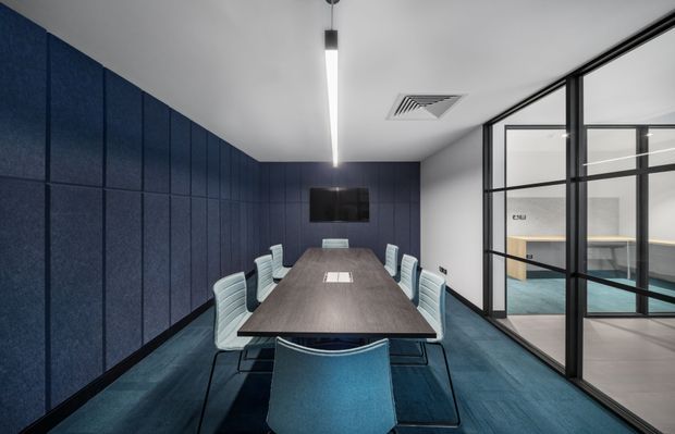 A contemporary meeting room with a long dark wood conference table, pale upholstered chairs, deep blue acoustic wall panelling and blue carpet. A linear pendant light provides focused illumination, while black-framed glazed partitions reveal an adjacent workspace. The room has a clean, professional and slightly moody corporate interior.