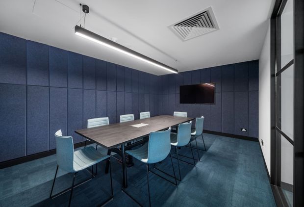 A compact, contemporary meeting room with a long dark wood meeting table surrounded by pale blue upholstered chairs. The walls are lined with dark navy acoustic panels, complemented by blue-toned carpet flooring. A wall-mounted screen, linear pendant light, exposed ceiling air vent, and black-framed glazed partition complete the clean commercial fit-out.