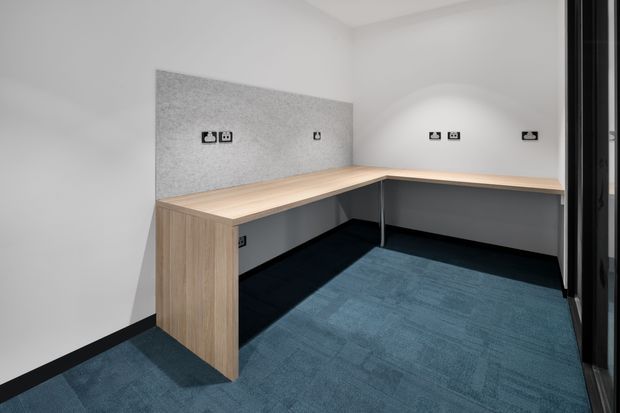 A compact, modern office workspace with an L-shaped built-in timber-look desk, grey acoustic wall panels, dark skirting, and blue carpet flooring. Multiple power and data outlets are integrated into the walls, with glazed or reflective partitions visible at the right. The space is clean, functional, and minimally furnished.