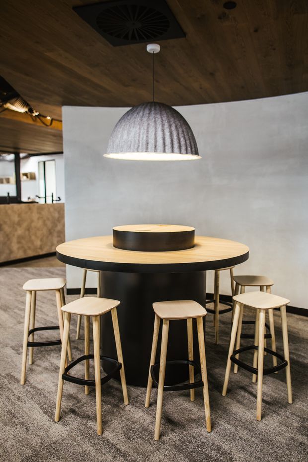 A compact contemporary dining or breakout area featuring a round light-timber and black pedestal table surrounded by tall timber stools. The space has a dark timber ceiling, grey carpet, a textured grey pendant light, a pale curved wall, and a partially visible kitchenette or service area in the background.