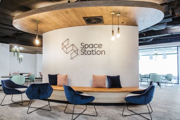 A contemporary commercial workspace reception or lounge area featuring a curved built-in upholstered bench, deep blue and blush lounge chairs, and a large rounded timber ceiling feature. A curved white partition wall displays the “Space Station” logo, while pendant lights, exposed services, and glazed meeting or work areas extend behind it.