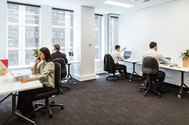 Bright contemporary open-plan office with several individual workstations, white desks, ergonomic black office chairs, computers, large windows, white walls, ceiling tiles and recessed lighting. The space has dark carpet flooring and a clean, professional commercial fit-out.
