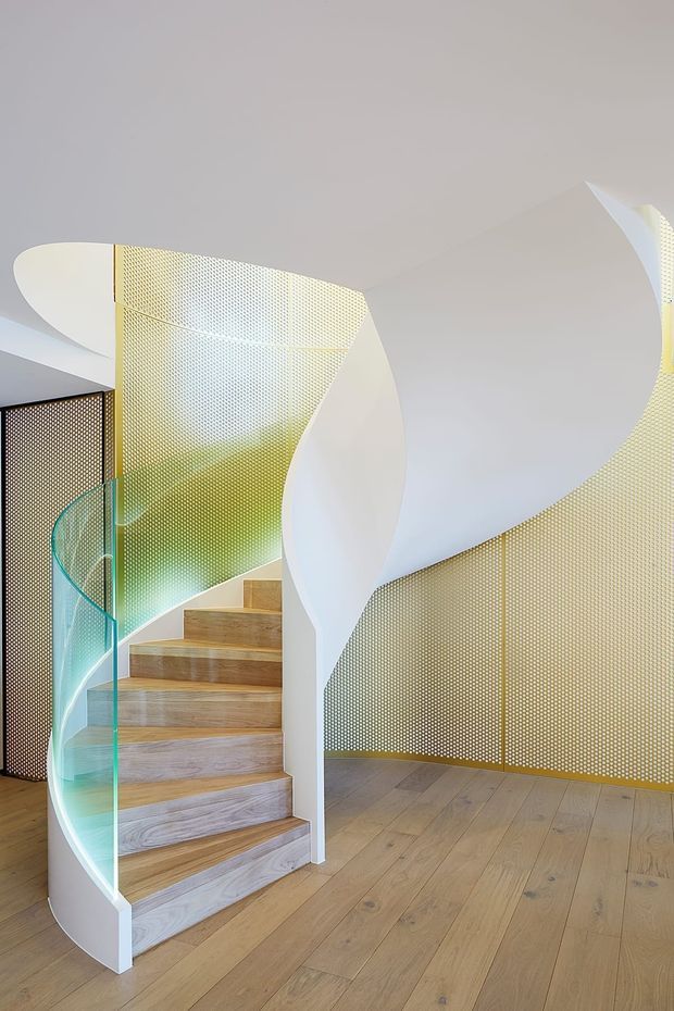 A sculptural contemporary stairway with pale timber treads and flooring, a sweeping white curved stair stringer and balustrade, and a clear curved glass guardrail. Perforated golden metal screens wrap the stair and upper wall areas, creating a distinctive patterned architectural feature. The bright, open composition focuses on the staircase and surrounding landing.