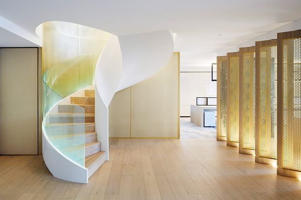 A bright, refined contemporary interior centred on a sculptural curved staircase. The staircase has pale timber treads, a translucent curved glass balustrade and a sweeping white sculptural side wall. Perforated gold-toned metal screens create a patterned divider along the right side, with pale timber flooring, white walls and glimpses of minimalist cabinetry beyond.