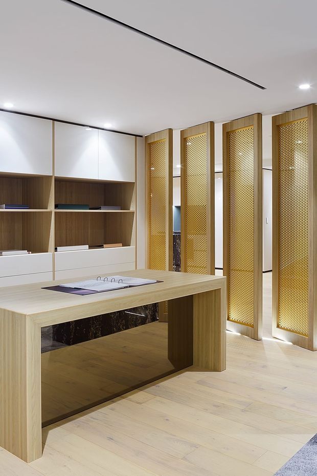 A bright, refined home office with extensive light timber built-in cabinetry, open shelving, and white upper cupboards. A large timber worktable with an open base occupies the foreground, while a series of tall timber-framed decorative screens with fine gold-toned mesh divide the workspace from the adjoining area. Pale timber flooring, recessed ceiling lighting, and dark trim details complete the contemporary fit-out.