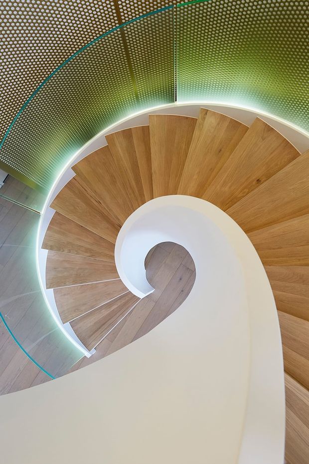 Top-down view of a sculptural curved spiral staircase. Warm timber treads wind around a central opening, bordered by a smooth white balustrade or stair structure. The surrounding wall and ceiling are covered in perforated green panels, with concealed linear lighting tracing the stair curve and turquoise edge detailing. The image emphasizes the dramatic geometry, craftsmanship, and contrast between timber, white surfaces, and green perforated panels.