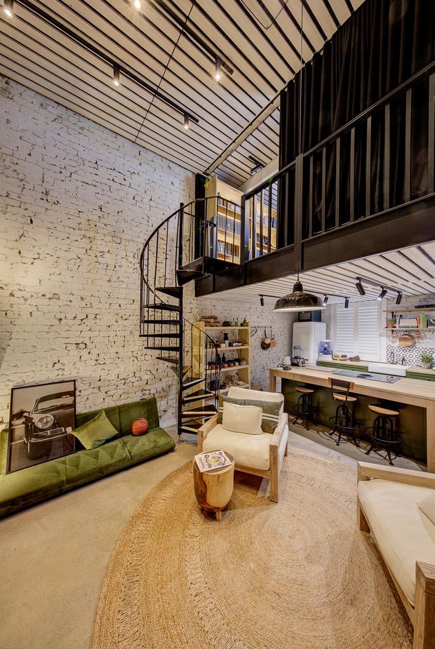 A loft-style open-plan living space with a kitchen beneath a black metal mezzanine. The room features exposed white-painted brick walls, a high slatted ceiling with track and pendant lighting, a black spiral staircase, dark metal-framed glazing and balustrades, timber kitchen cabinetry, bar seating, and relaxed lounge furniture arranged on a large natural-fibre rug.