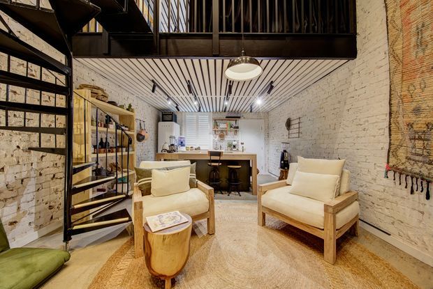 A warm, industrial-style open-plan living area in a converted brick interior. The room features exposed white-painted brick walls, a black steel spiral staircase and mezzanine balustrade, open black metal shelving, timber lounge chairs, a rounded timber side table, a compact kitchen or bar counter, and a slatted ceiling with pendant and recessed lighting.