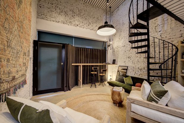 A loft-style living room with exposed white-painted brick walls, a black metal spiral staircase, and a small bar-height counter with stools. The room features a slatted timber ceiling, dark-framed glazed doors with curtains, pendant lighting, a pale area rug, and light timber-framed seating with white and olive cushions. The overall atmosphere is warm, relaxed, and industrial-modern.