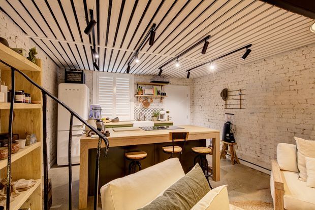 A warm, compact open-plan living and kitchen area in a converted brick interior. The room features exposed painted brick walls, a slatted timber ceiling with black track lighting, timber open shelving, a central kitchen island with timber benchtop and stools, white cabinetry and appliances, and a partially visible sofa in the foreground. A black metal stair railing and steps are visible at the left.