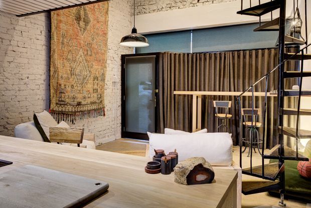 An industrial-style open-plan living and dining space with white-painted brick walls, exposed timber ceiling slats, a large woven wall hanging, black-framed glazing, a timber dining table, and a black metal spiral staircase leading to an upper level. A curtained sleeping or living area is visible behind the dining zone.