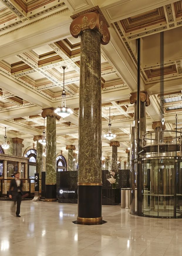 Grand ornate commercial banking or office lobby with monumental green-marble columns, bronze and gold Corinthian-style capitals, highly decorated coffered ceilings with geometric Greek-key detailing, polished stone flooring, chandeliers, arched windows, and a cylindrical glass elevator enclosure. The space has a heritage-inspired, opulent character with contemporary reception and security elements.