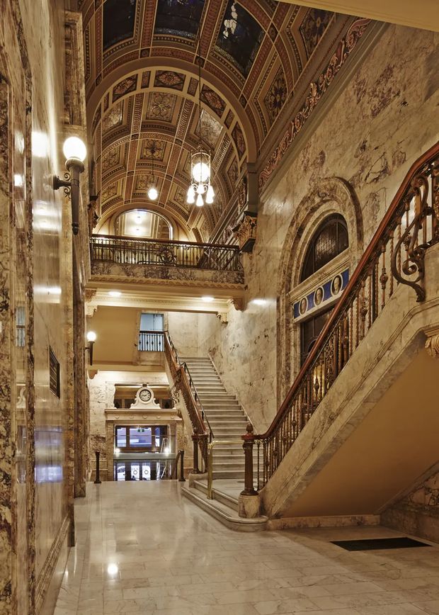 Grand historic interior hall with a polished marble floor and marble-clad walls, an ornate bifurcated staircase with dark timber and decorative metal balustrades, upper-level gallery, arched openings, and a richly patterned barrel-vaulted ceiling. Warm globe and chandelier lighting emphasize the formal heritage character of the space.