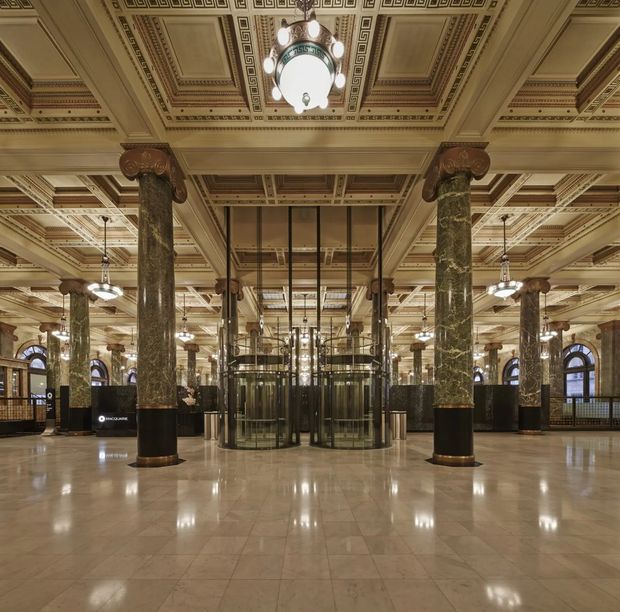 Grand historic commercial interior resembling a bank or civic hall, with a symmetrical open lobby, ornate coffered and patterned ceiling, monumental green marble columns with decorative capitals, polished stone flooring, arched windows, and central glass revolving doors or lift enclosures. The space has a richly detailed classical architectural character and warm subdued lighting.
