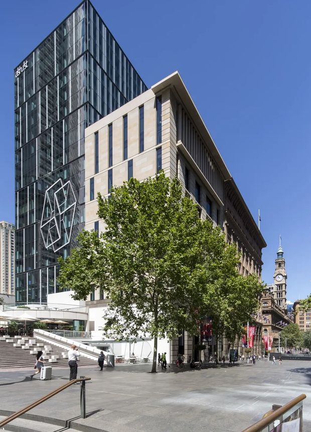 Street-level exterior view of a contemporary multi-storey commercial building complex in a prominent urban civic plaza. The main building combines a tall glazed curtain-wall tower with a lower stone-clad wing featuring vertical window bays, deep roof overhangs, broad steps, and public plaza paving. Mature trees, pedestrians, outdoor umbrellas, and a historic clock tower in the background reinforce the dense city-centre setting.