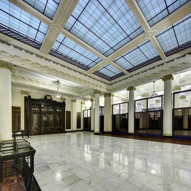 A grand historic interior hall with a large glazed roof supported by ornate cream-painted beams, elaborate classical cornices, decorative column capitals, and tall cylindrical columns. The space has a highly polished pale stone or marble floor, dark timber and glazed partitioning, ornate dark entrance doors, period pendant lights, and formal architectural detailing. The image appears to show a heritage public or commercial building lobby rather than a residential room.