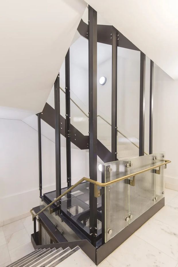 A bright contemporary stairwell with a multi-level staircase enclosed by transparent ribbed glass balustrades and dark metal structural frames. Brass-toned handrails provide contrast, while white walls, ceilings and pale stone or tile flooring create a clean, open vertical circulation space. Circular wall lights and glimpses of lower stairs are visible.