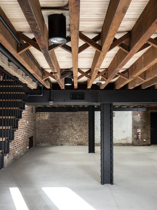 An open-plan industrial interior with exposed timber roof trusses and beams, black-painted steel structural columns and ductwork, exposed aged brick walls, a pale polished concrete floor, and an industrial black metal stair or shelving structure along the left wall. The space is largely empty and lit by overhead fixtures and daylight.