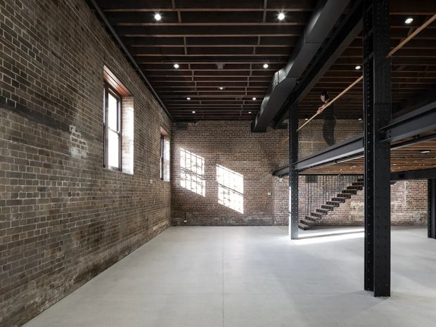 An expansive adaptive-reuse interior with exposed aged brick walls, a smooth concrete floor, and a dark exposed structural steel mezzanine. Open-riser stairs lead to the upper level, while large industrial windows and exposed ceiling framing bring in daylight. The space is largely empty and warehouse-like.