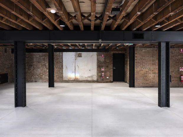 A raw industrial interior with an open concrete floor, exposed brick walls, heavy black steel columns and beams, and an exposed timber joist ceiling. The space appears unfinished or adaptively reused, with minimal fixtures and service elements visible.