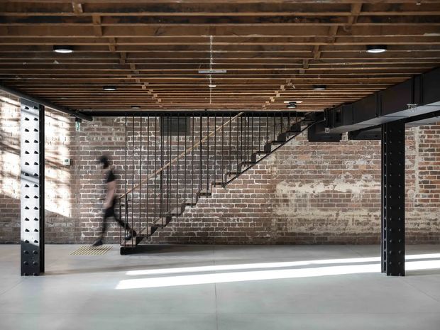 An open-plan industrial-style interior with exposed red brick walls, a dark steel structural frame, and a black metal staircase with slender balustrades. The ceiling features exposed timber joists and services, while the polished light-grey concrete floor is crossed by strong sunlight. The space appears largely unoccupied and could function as a converted warehouse or commercial workspace.