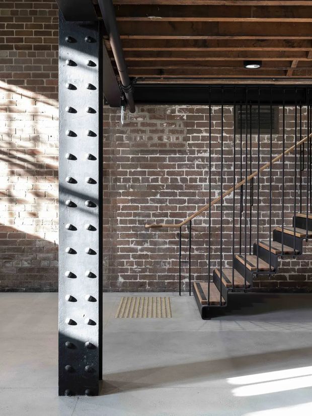 Industrial-style interior featuring an exposed red-brick wall, raw timber ceiling joists, a perforated steel structural column, and a black metal staircase with timber treads and railing. The concrete floor and exposed services reinforce the utilitarian character, while daylight creates strong diagonal shadows across the space.