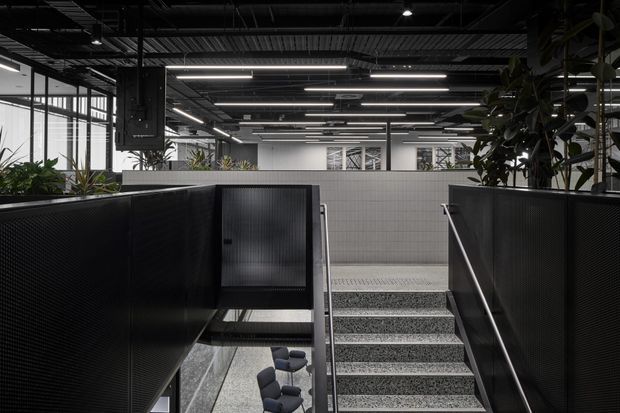 A modern commercial interior viewed from a stair landing, showing broad terrazzo-finished stairs with metal handrails, black perforated privacy partitions, tiled walls, exposed dark ceiling services, linear strip lighting, and abundant indoor planting. Open-plan office work areas are visible beyond the stairwell.