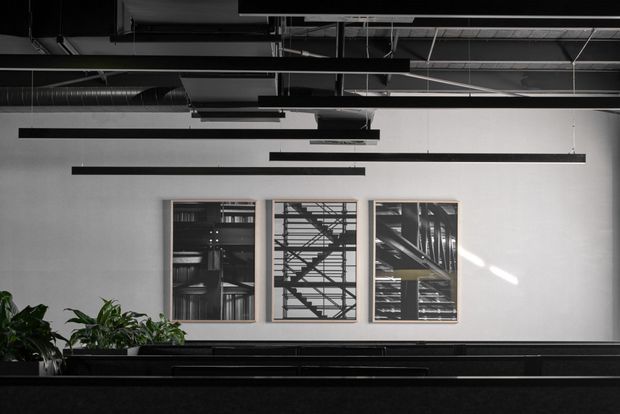 A monochrome commercial interior featuring an exposed industrial ceiling with ducts, suspended linear lights, and black structural elements. A long dark reception or counter runs across the foreground, with plants at the left and three framed architectural or structural images mounted on a pale wall.