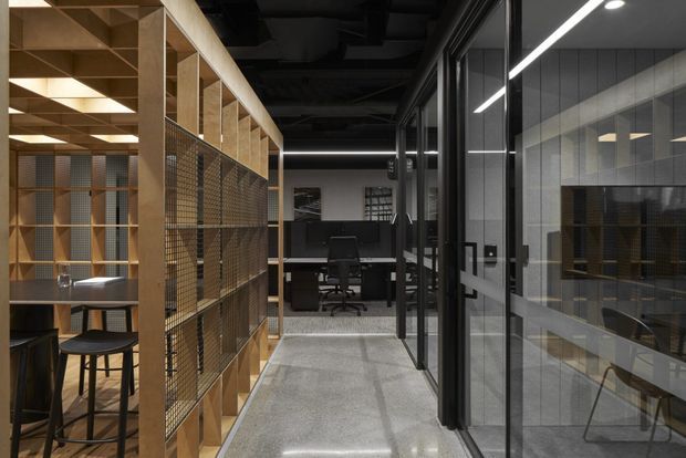 A contemporary commercial office interior with a polished concrete corridor between glass-partitioned rooms and a timber-framed open workspace. The space features black ceilings, linear lighting, metal mesh screens, built-in timber shelving, workstations, and dark office furniture.