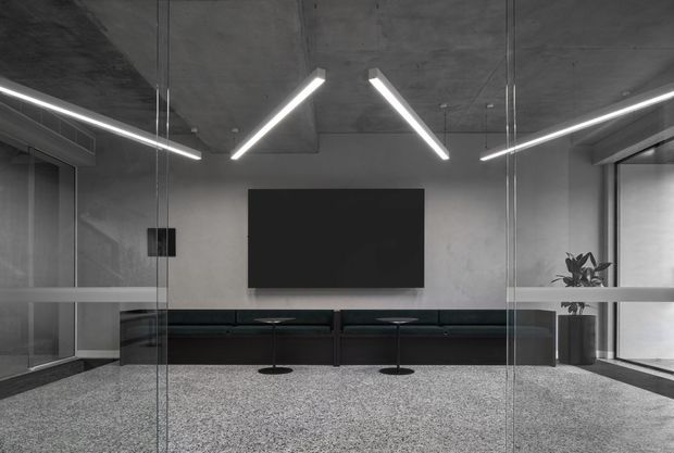 A sleek minimalist commercial interior viewed through full-height glass partitions. The space features a raw concrete ceiling and walls, linear suspended LED lighting, a large black wall-mounted display, built-in dark seating, a speckled terrazzo floor, and a potted plant. The monochrome palette and exposed finishes create a refined, contemporary office or media area.