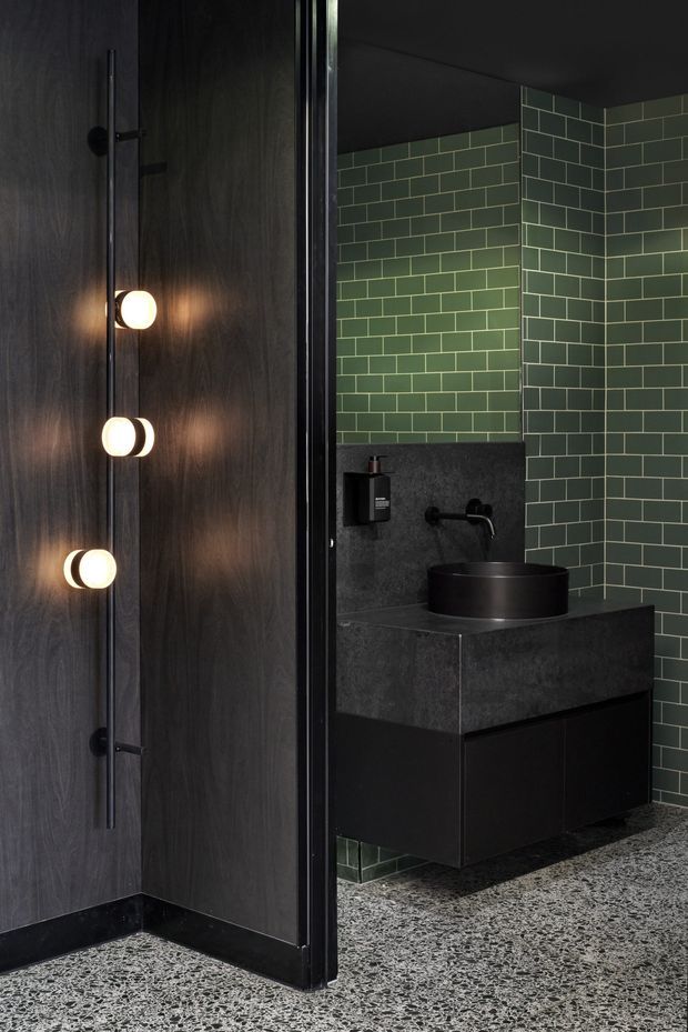 A dark, moody bathroom viewed through a partially open doorway. The room features deep green rectangular wall tiles, a black vessel basin on a charcoal stone vanity with black cabinetry, a wall-mounted black tap, dark timber wall panelling, a terrazzo floor, and a tall sculptural wall light on the door.
