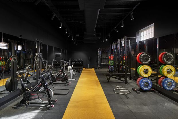 A spacious commercial-style gym with black walls and exposed dark ceiling services, rows of cardio and strength-training equipment, weight racks with brightly coloured plates, track lighting, and a bold yellow central rubber walkway.