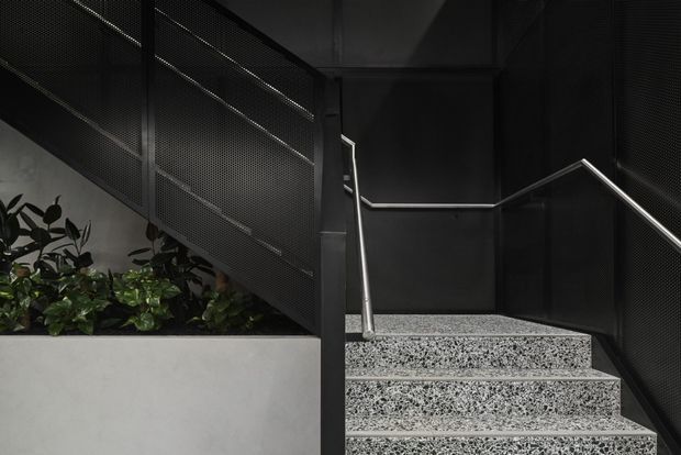 A dark, modern stairwell with exposed speckled concrete stairs, stainless-steel handrails, black perforated metal balustrade and wall panels, and a built-in planter containing lush greenery. The composition is architectural and industrial, with strong geometric lines and subdued lighting.