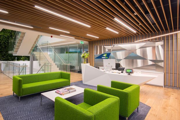 A bright contemporary commercial reception and waiting area with a white angular reception desk, green upholstered sofas and armchairs, pale timber flooring, and a dark patterned carpet. A dramatic slatted timber ceiling with integrated linear lighting extends across the space, while glass balustrades and partitions reveal a stairway and additional circulation areas. The reception backdrop features a faceted metallic geometric wall.