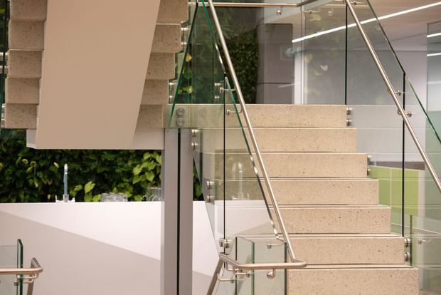Contemporary interior stairway with broad terrazzo or aggregate-finished steps, clear glass balustrades, and slim brushed metal handrails. A sculptural white stair structure and partial white cabinetry or partitioning frame the scene, with a lush green living wall visible behind. Bright linear lighting and clean geometric detailing create a refined modern commercial or residential setting.