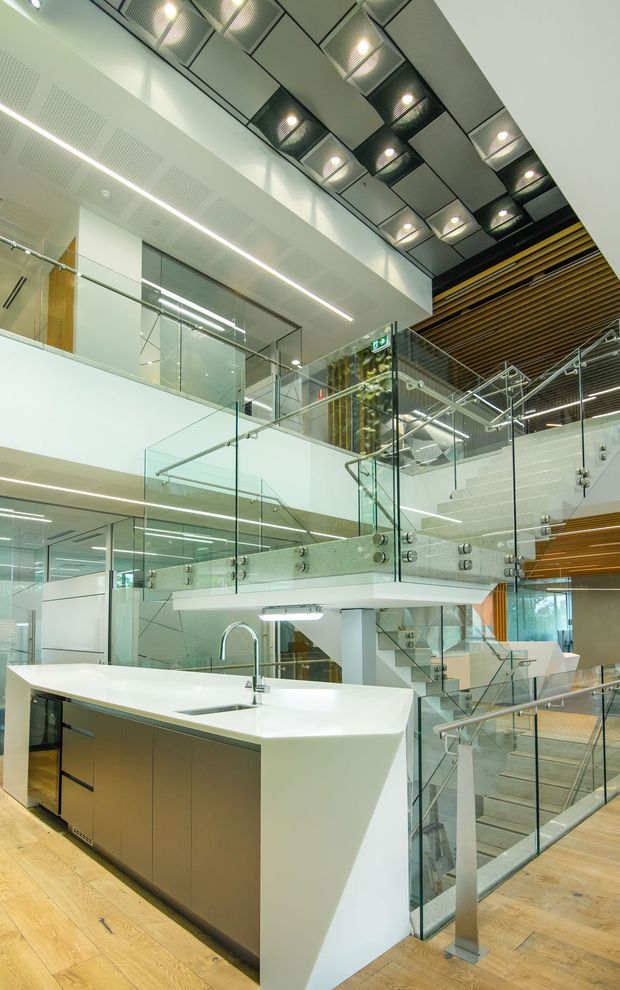 A bright, multi-level commercial interior with a contemporary open stair and circulation space. The foreground features a large white island kitchenette or service counter with warm wood cabinetry and a chrome gooseneck tap. Multiple stair flights and mezzanine edges are enclosed by clear glass balustrades with stainless-steel fixings. The space has white walls, extensive linear lighting, exposed acoustic ceiling panels, and a striking ceiling installation of recessed lights in black-framed geometric panels. Warm timber slatted ceiling elements and timber flooring add contrast to the predominantly white and glass interior.