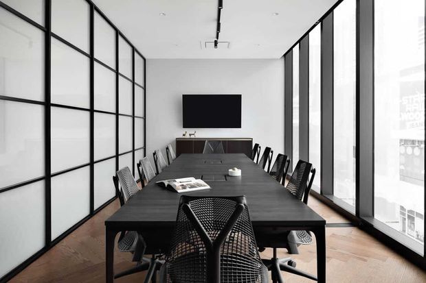 A contemporary meeting or dining room with a long dark table surrounded by mesh-backed chairs. Black-framed translucent glass partitions line the left wall, while full-height glazing runs along the right side. A wall-mounted black screen and low dark credenza sit at the far end. The room has white walls and ceiling, dark timber flooring, linear track lighting, and a restrained monochromatic palette.