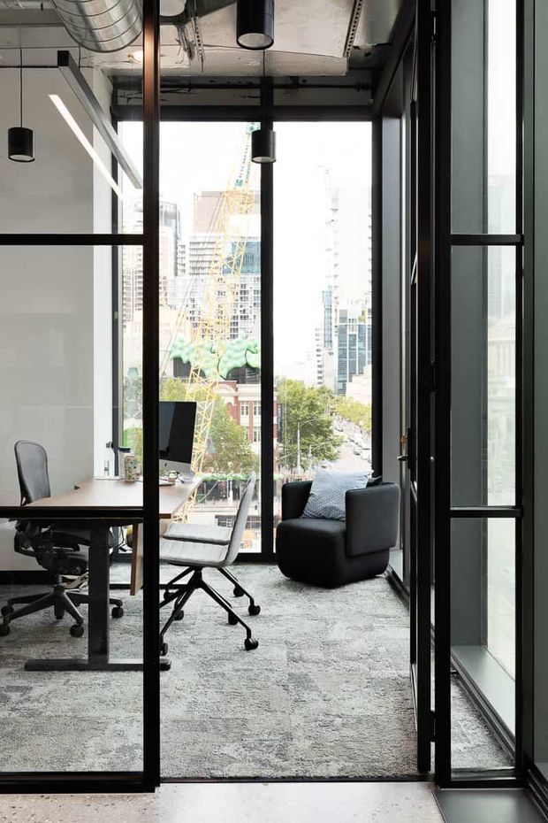 A modern urban office interior viewed through black-framed glazed partitions. The space features exposed industrial-style services and ductwork, concrete ceiling surfaces, floor-to-ceiling windows, carpet flooring, a timber desk with office chairs, and a dark lounge armchair. High-rise buildings and construction activity are visible outside.